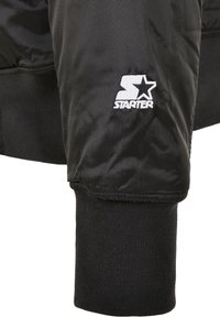 Giubbotto piumino nero con polsini a coste e logo ricamato bianco "Starter" con una stella. Il materiale ha una finitura liscia e lucida.