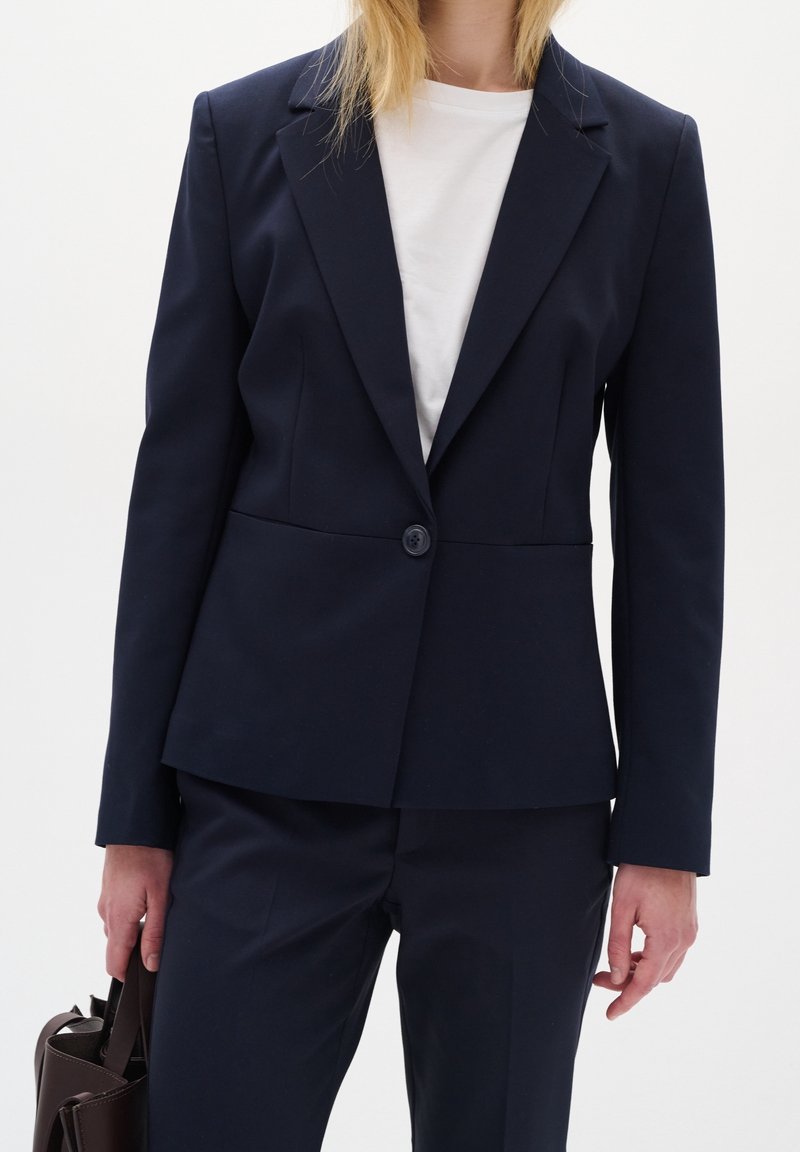 Personne portant un blazer bleu marine ajusté et un pantalon assorti avec une chemise blanche, tenant un sac à main marron foncé sur un fond clair.