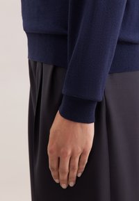 Maglione a collo alto di colore blu navy con polsini a costine, abbinato a una gonna a pieghe scura. Primo piano del braccio e della mano, che mette in evidenza la texture del tessuto.