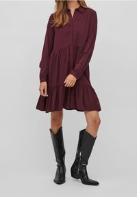 Abito camicia bordeaux con maniche lunghe, pattina dei bottoni, gonna a strati, indossato con stivali neri al ginocchio con dettaglio zip.