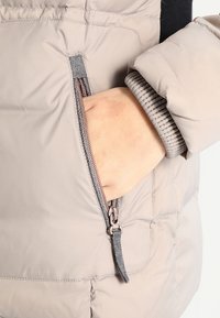 Mano dentro del bolsillo con cremallera de una chaqueta de invierno acolchada de color beige claro con puños elásticos.