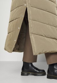 Abrigo acolchado beige con textura acolchada y cierre de botones a presión. Llevado sobre pantalones marrones ajustados y botas chelsea negras con costuras amarillas.