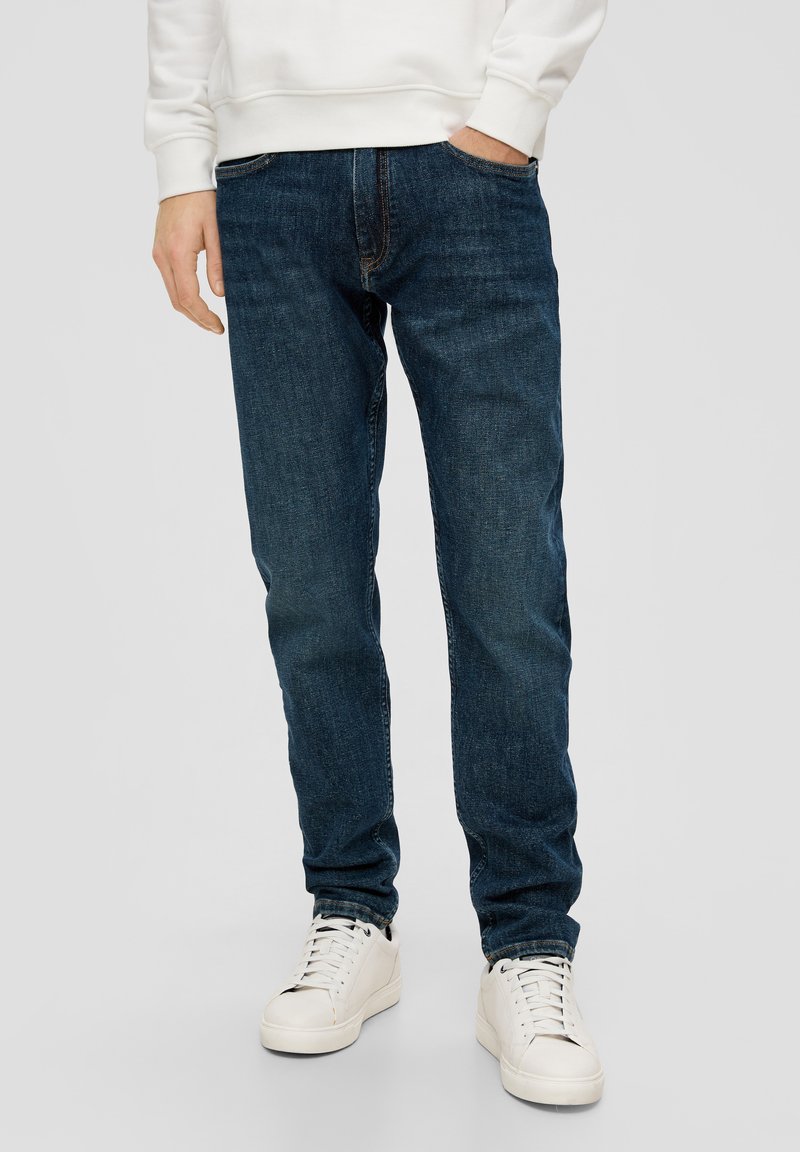 Jean en denim bleu foncé avec une coupe droite, cinq poches, une couture discrète et une texture usée, associés à des baskets blanches.