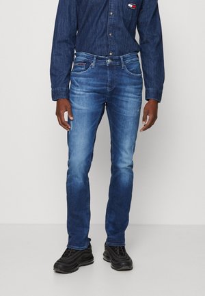 Hombre con vaqueros azules descoloridos, camisa vaquera azul oscuro con logotipo en el bolsillo del pecho y zapatos deportivos negros de pie sobre un suelo liso.