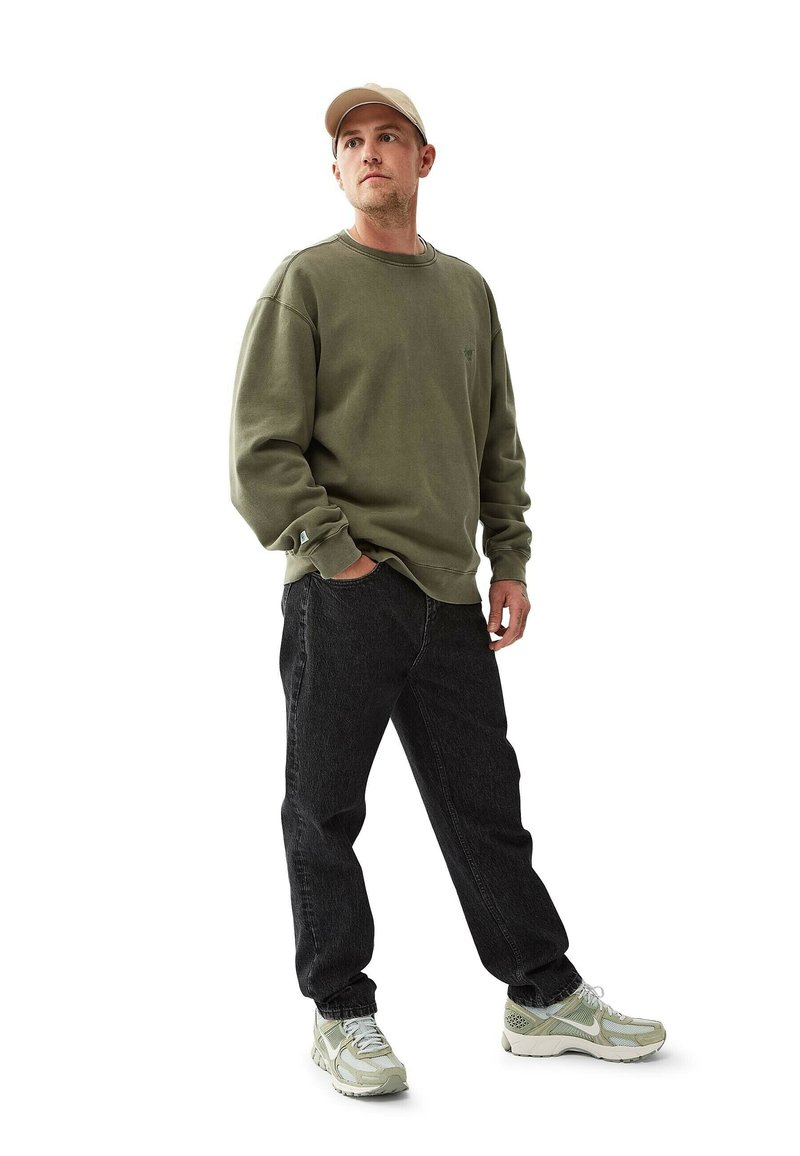 Man i beige keps, olivgrön sweatshirt, svarta jeans och vita sneakers, stående med en hand i fickan mot vit bakgrund.