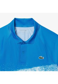 Sininen polo-paita, jossa on kaulus, kaksinappinen etuosa ja vihreä krokotiililogo. Kangas on sileä ja siinä on gradienttimuotoilu.