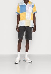 Camisa de manga corta multicolor con un diseño de patchwork, que presenta patrones en azul, amarillo y blanco; combinada con pantalones cortos de mezclilla oscura y zapatillas.