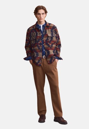 Joven de pie con las manos en los bolsillos, vestido con una camisa de botones con estampado, camiseta azul, pantalones marrones y zapatos oscuros.