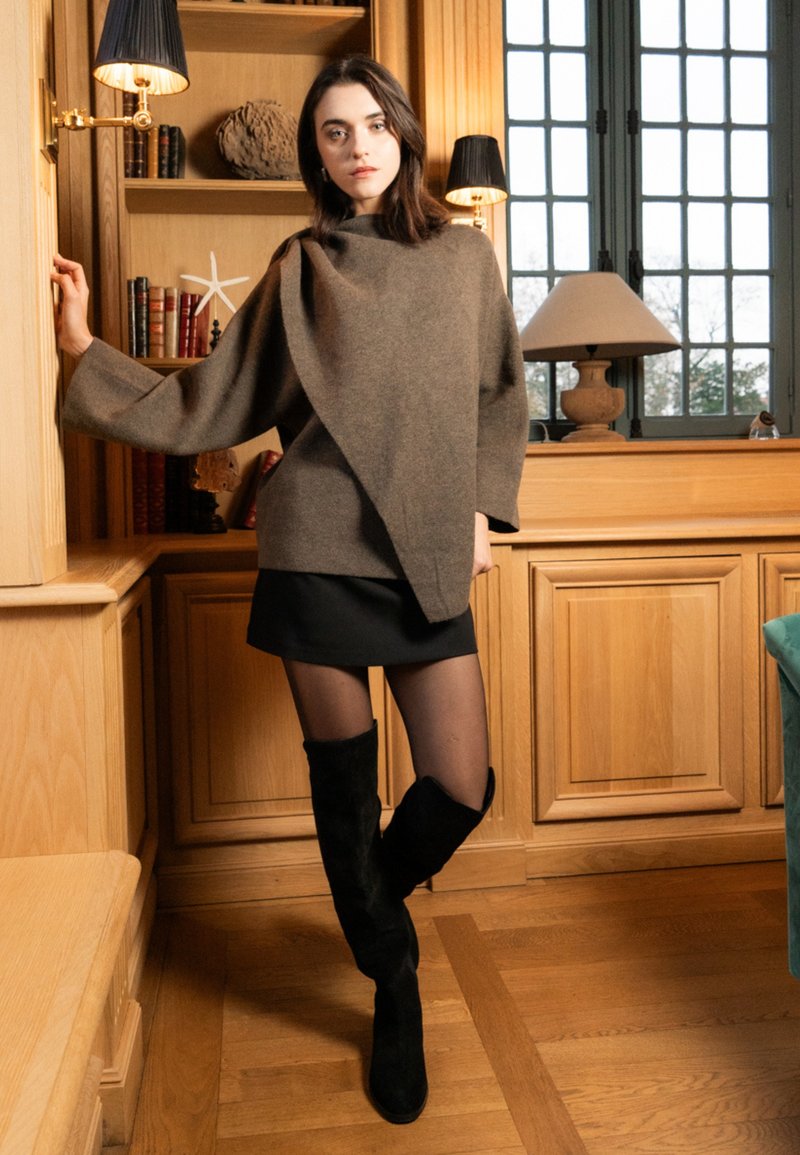 Brązowa wełniana bluzka typu wrap, czarna mini spódnica, przeźroczyste rajstopy i wysokie czarne zamszowe kozaki. Modelka stoi obok drewnianych szafek i lamp.