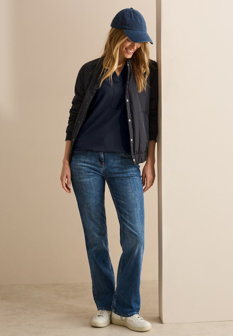 Veste bomber matelassée noire sur un polo bleu marine, assortie à un jean flare bleu et des baskets blanches. Une casquette bleue complète le look décontracté.