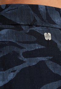 Pantalones cortos en color marino con un patrón texturizado en diferentes tonos de azul, logotipo de herrajes plateados y un acabado suave en la cintura.