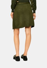 Persona che indossa una gonna verde oliva e un maglione abbinato con mocassini neri, in piedi con le mani rilassate lungo i fianchi, spalle rivolte verso la macchina fotografica.