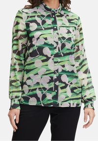 Donna che indossa una camicia con bottoni a strisce astratte verdi e nere con motivi floreali bianchi, abbinata a pantaloni neri.