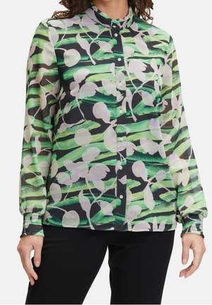 Femme portant une blouse à boutons avec des rayures abstraites vertes et noires et des motifs floraux blancs, assortie à un pantalon noir.