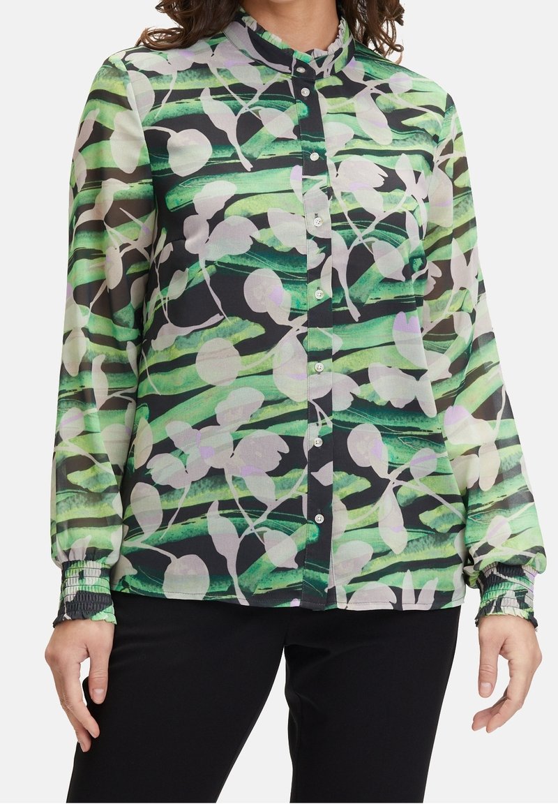 Donna che indossa una camicia con bottoni a strisce astratte verdi e nere con motivi floreali bianchi, abbinata a pantaloni neri.