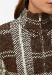 Adīta d�žemperis ar augstu apkakli un pusceļam, brūnā un krēmkrāsā ar ģeometriskām rakstām un faktūras detaļām.