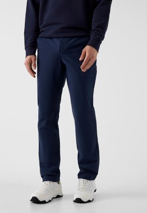 Hombre con pantalones azul marino de corte recto, zapatillas deportivas blancas y una camisa azul marino de manga larga sobre un fondo gris liso.