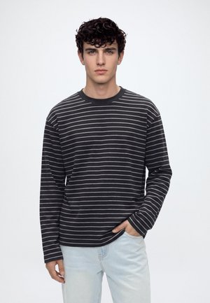 Jeune homme aux cheveux bouclés foncés portant un t-shirt à manches longues rayé noir et blanc et un jean bleu clair, debout avec une main dans la poche.