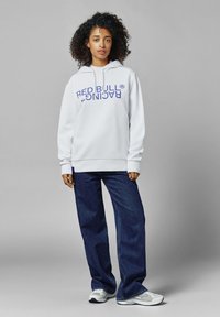Sudadera blanca con el texto "RED BULL RACING" en azul, combinada con jeans de pierna ancha azul oscuro y zapatillas grises. Tejido suave y corte relajado.