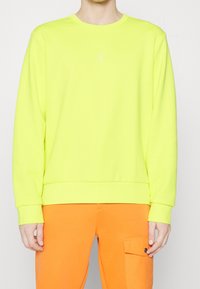 Neon žlutá mikina s kulatým výstřihem a dlouhými rukávy, s žebrovaným lemem. Nositelná s oranžovými kalhotami, které mají postranní kapsu.