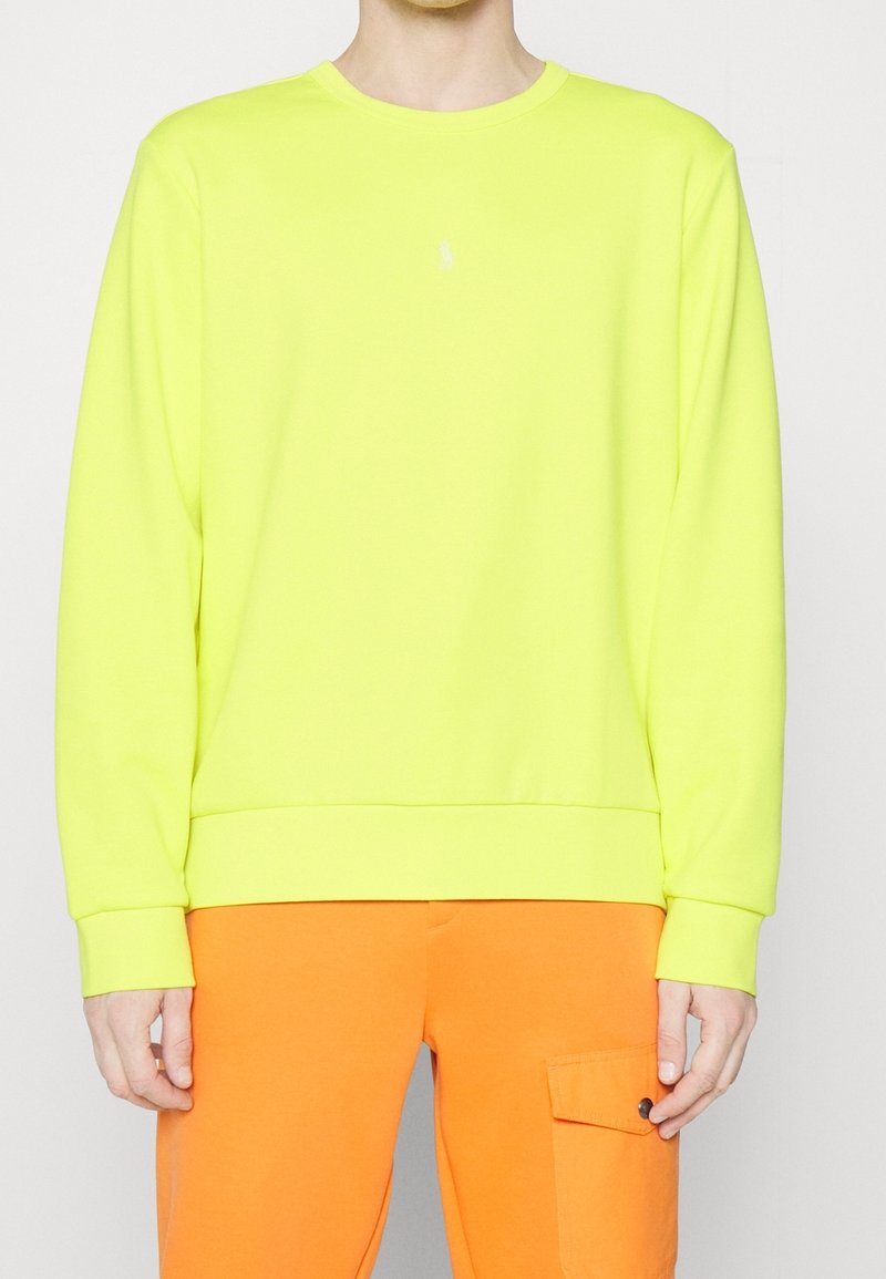 Neon žlutá mikina s kulatým výstřihem a dlouhými rukávy, s žebrovaným lemem. Nositelná s oranžovými kalhotami, které mají postranní kapsu.
