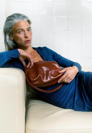 Borsa in pelle marrone dalla forma arrotondata, appoggiata contro un vestito blu texturizzato, posata su un divano in pelle bianca. Design semplice, senza elementi di hardware prominenti.