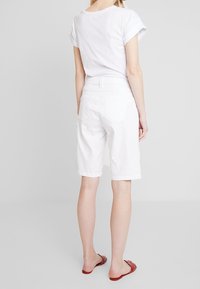 T-shirt en coton blanc avec manches retroussées, associé à un short blanc à mi-genoux avec une fermeture éclair à l'avant et deux poches arrière. Sandales plates rouges.