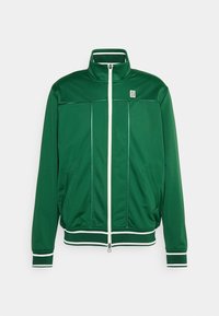 Groene ritsjack gemaakt van een gladde stof. Kenmerkt zich door een hoge kraag, witte ritsen en contrasterende afwerking rond de manchetten en de zoom.