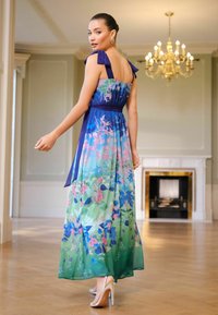 Kwiatowa sukienka maxi w niebiesko-zielonym gradiencie, z kokardami na ramionach, dopasowaną talią oraz zwiewną spódnicą o żywych wzorach. Srebrne szpilki.