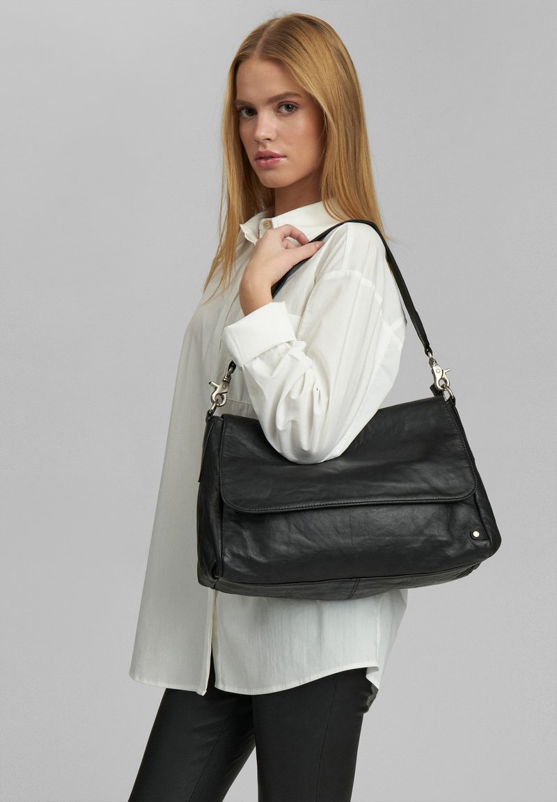 Borsa a spalla nera in pelle con chiusura a pattina, hardware argentato, superficie texturizzata e tracolla regolabile. Abbinata a una camicia bianca.
