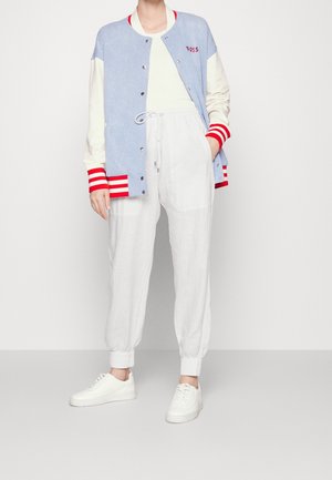 Osoba ubrana w jasnoniebieską i kremową kurtkę varsity z czerwonymi paskami, białe spodnie z troczkami oraz białe trampki, stojąca z jedną ręką w kieszeni.