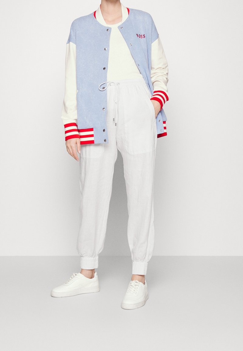 Personne portant une veste universitaire bleu clair et crème avec des rayures rouges, un pantalon blanc à cordon de serrage et des baskets blanches, debout avec une main dans la poche.