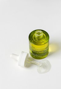 Botella de vidrio verde con un tapón gotero blanco, que contiene un líquido transparente; una gota del líquido reposa sobre una superficie blanca.