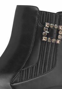 Botte cheville en cuir noir présentant une texture à nervures verticales et une boucle décorative ornée de gemmes transparentes et fumées sur le côté.