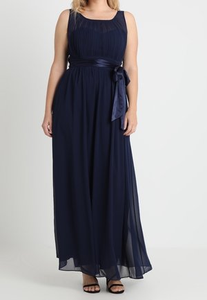 Woman wearing a sleeveless navy blue pleated floor-length dress with a satin waist bow and black open-toe heels, standing against a white background.