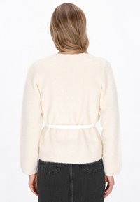 Cream-colored, fluffy sweater with long sleeves and a fitted waist, secured by a white belt. Soft texture, round neckline, open back design.