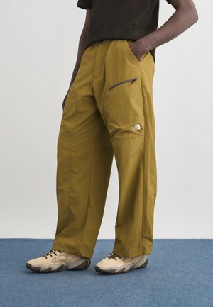 Modello che indossa pantaloni da trekking giallo senape a gamba larga con tasca laterale con zip e scarpe beige da trail su pavimento con moquette blu.