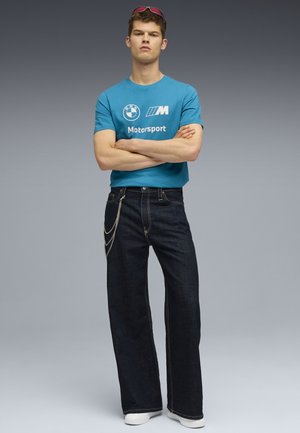 Teal bomuld t-shirt med BMW Motorsport logo, parret med mørke denim bukser med brede ben, der har en sølv kædeaccent. Hvide sneakers fuldender looket.