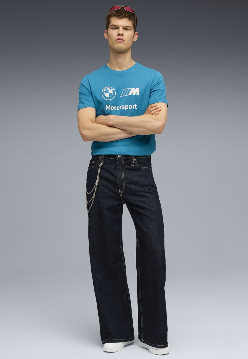 Mėlynas medvilninis marškinėlis su BMW Motorsport logotipu, derinamas su tamsiai mėlynomis plačiomis džinsinėmis kelnėmis, papuoštomis sidabrinėmis grandinėmis. Išvaizdą u�žbaigia baltos sportbačiai.