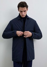 Hombre vistiendo un abrigo azul marino oscuro sobre un suéter de cuello alto negro, abrochándose el abrigo, de pie contra un fondo claro y liso.