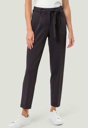 Frau trägt dunkelgraue, taillierte Hosen mit Bindegürtel und weiße Turnschuhe, steht vor einem schlichten hellen Hintergrund.