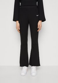 En person som bär svarta ribbade utsvängda byxor med en vit Nike-logotyp och vita sneakers, står mot en enfärgad bakgrund.