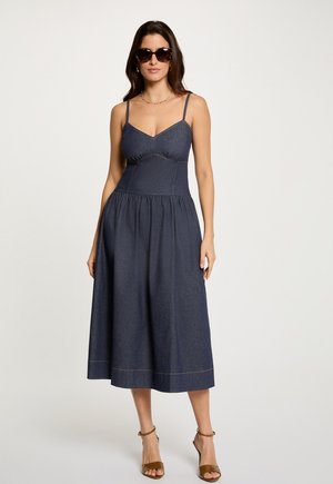 Femme portant une robe midi sans manches bleu foncé avec fines bretelles, des talons ouverts marron, des lunettes de soleil et des bijoux simples, debout devant un fond uni.