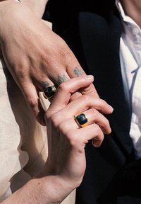 To hænder flettet sammen, hver iført en kraftig guldring med en mørk sten. Hudfarverne varierer, med detaljerede tatoveringer på den ene hånd.