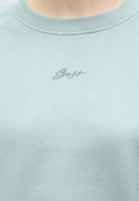Gros plan d'un sweat-shirt bleu clair à col rond avec un petit logo « Boss » brodé centré sous l'encolure.
