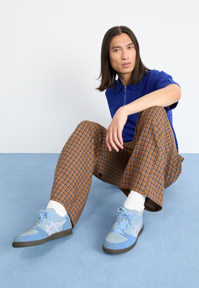Camisa polo texturizada azul, pantalones anchos de cuadros marrones, zapatillas de deporte azul claro con acentos grises y calcetines blancos, sentado contra un fondo claro.