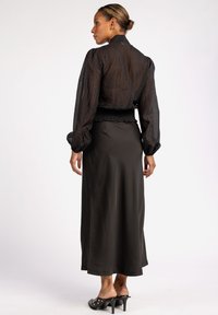 Zwarte doorschijnende blouse met lange mouwen, opgeblazen schouders en een smock taille, gecombineerd met een zwarte satijnen maxi rok en gestileerde, gestifte sandalen met hak.