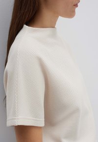 Top corto a maniche corte in maglia texturizzata di colore crema, dotato di un colletto alto e finiture sui polsini, che presenta un motivo sottile e un design strutturato.