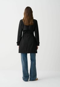 Černý trenčkot s přiléhavým korzetem, dlouhými rukávy a páskem v pase. Výrazný design zadního záhybu a rozparky. Kombinováno s rozšířenými modrými džíny.