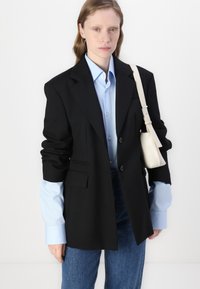 Črn krojen blazer z strukturiranimi rameni, z enim gumbom in sprednjimi žepi. Nošen preko svetlo modre srajce z zavihanimi rokavi.